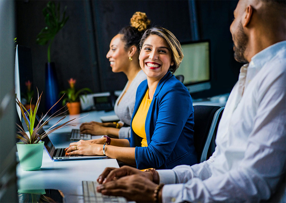 Three working professionals sitting at a desk smiling and working on laptop computers.